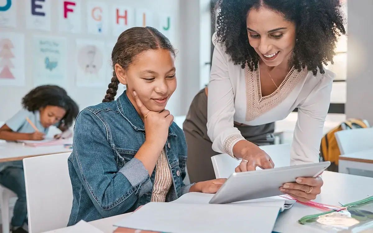 A teacher and a young student looking at a tablet together at a classroom desk
