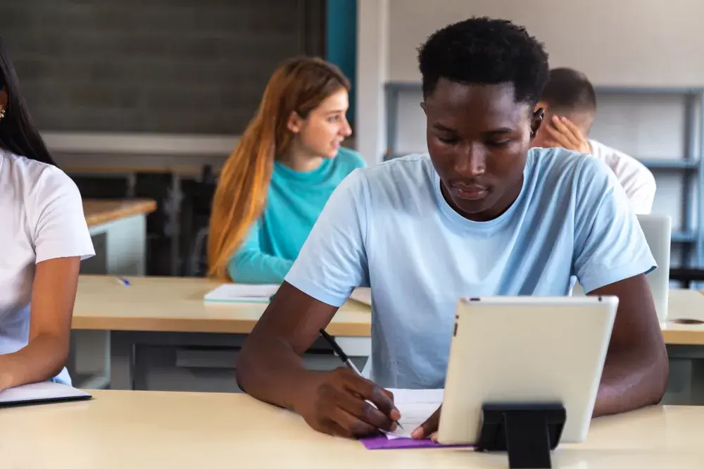 A college student takes notes by hand while working from a tablet at a classroom desk, with classmates seated behind.