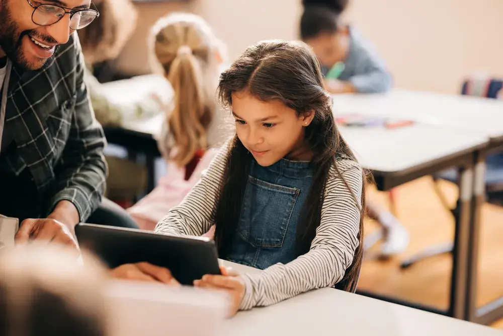 A student works on a tablet while an adult sits alongside, watching without touching the device.