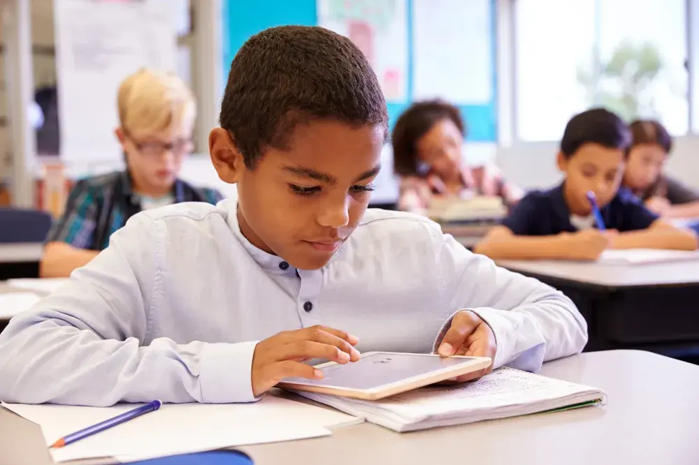 A student works through a session on a tablet at a classroom desk while classmates work nearby.