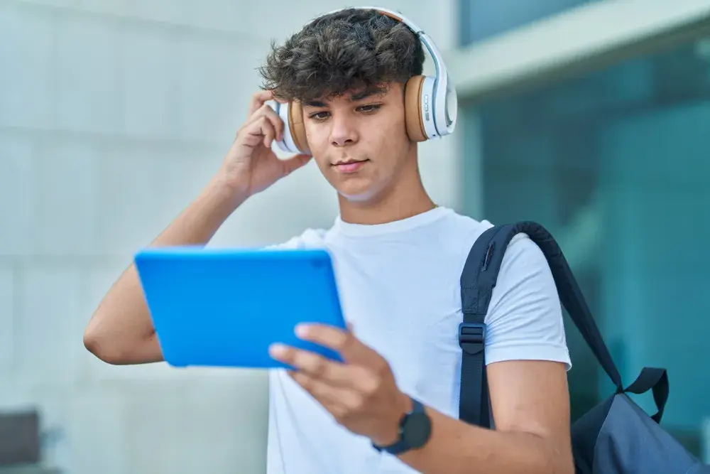 A student wearing headphones and a backpack uses a tablet outside school.