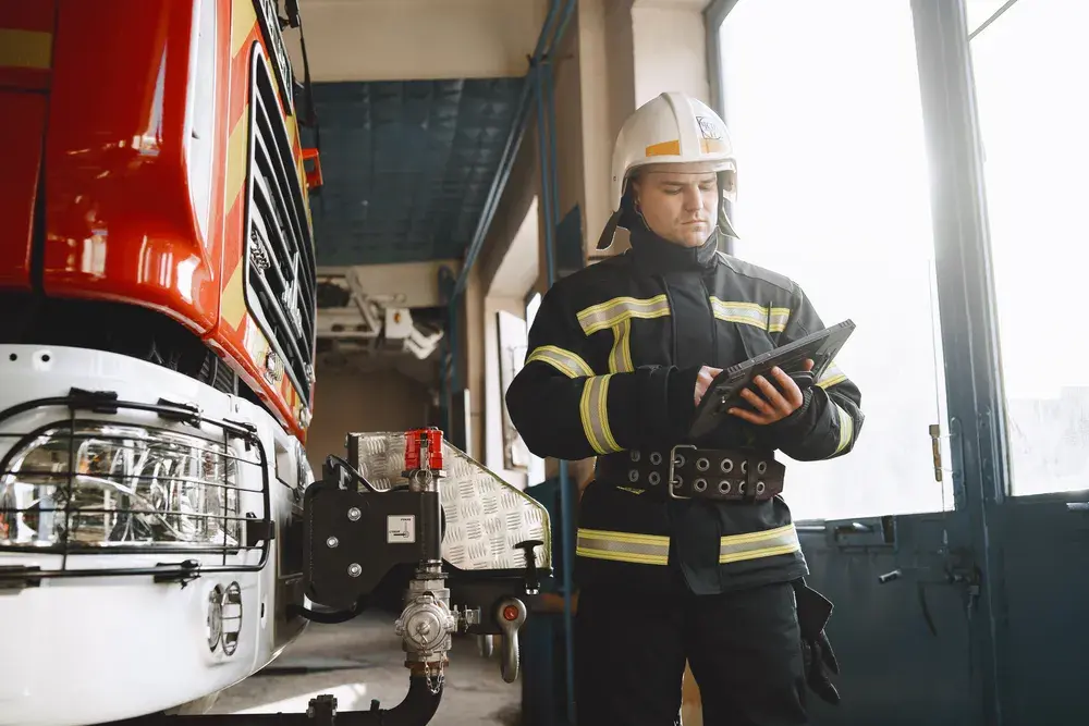 A firefighter in turnout gear and helmet using Mastermind on a tablet beside a fire engine in a station