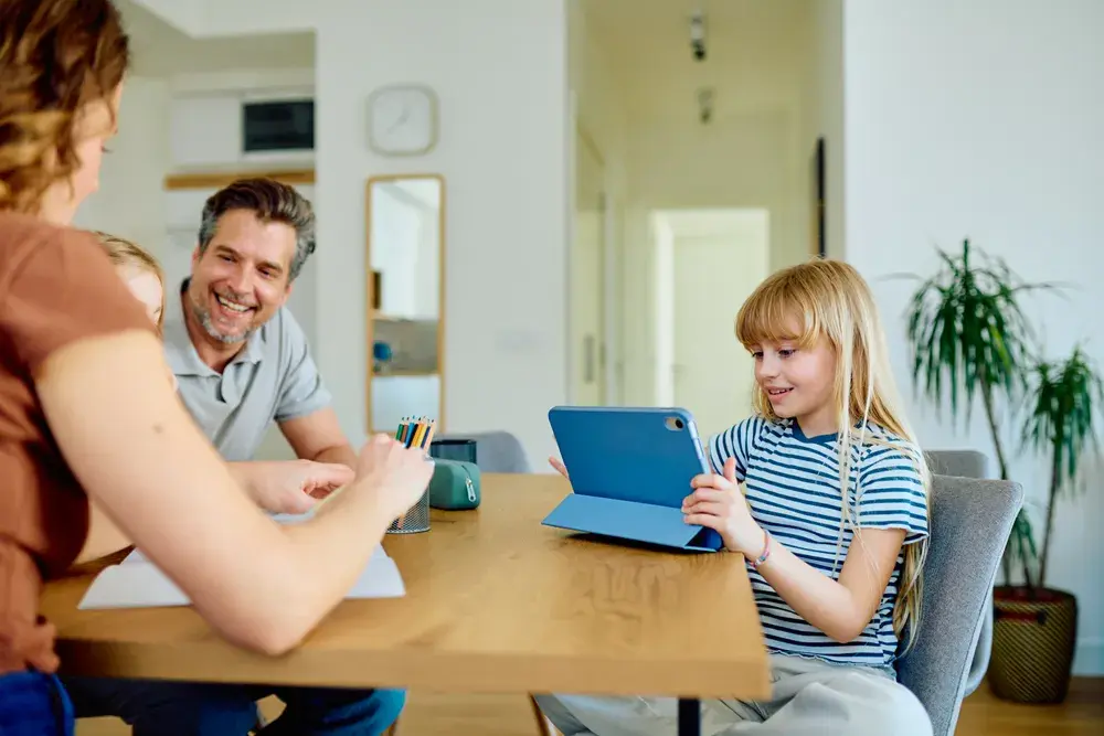 A girl wearing headphones works on a tablet at a table at home.