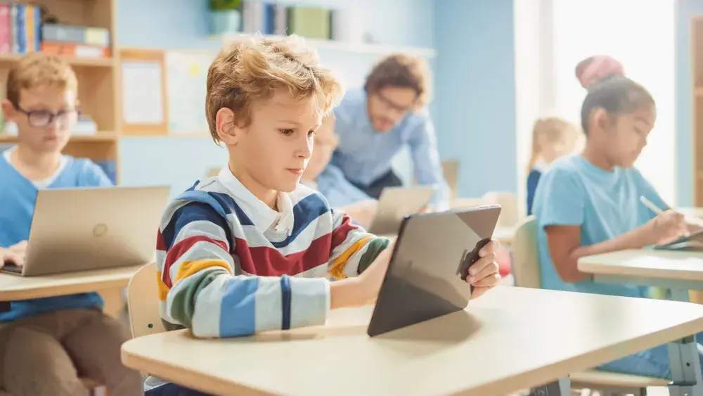 A high school student works through a session on a tablet at a classroom desk while a classmate works on separate material beside her.