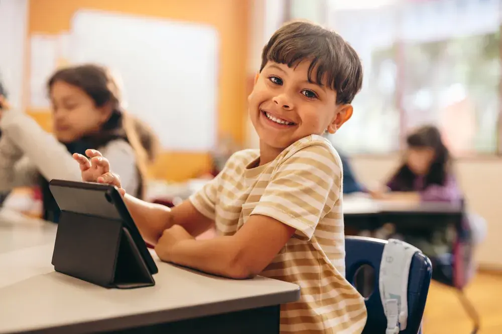 student smiling at camera, using a tablet in the classroom