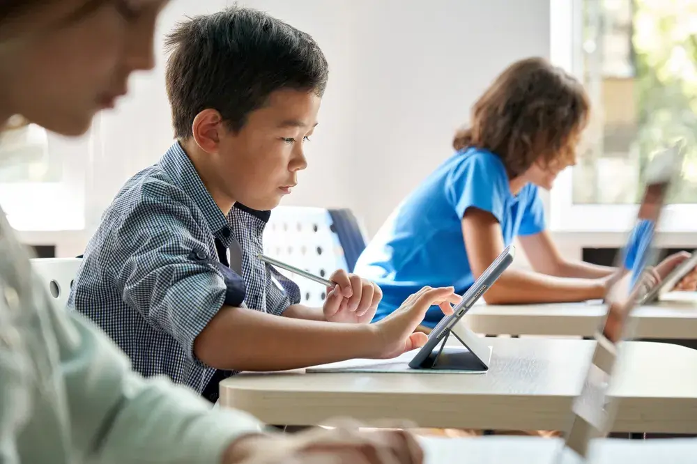 A student works through a session on a tablet at a classroom desk while classmates work nearby.